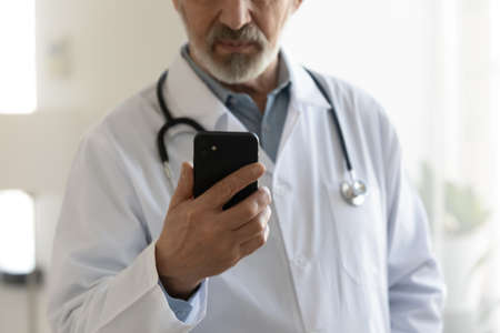 Close up focused mature doctor wearing white coat uniform with stethoscope holding smartphone,...