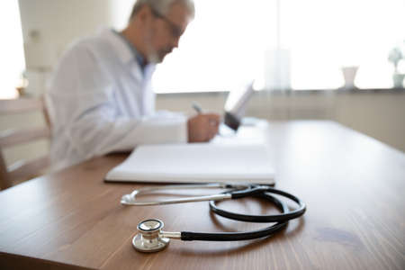 Close up senior mature doctor work desk with stethoscope, middle aged therapist practitioner gp sitting at table, writing in documents, filling illness history, patient card, working in hospitalの写真素材
