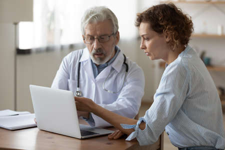 Serious mature doctor wearing glasses showing medical checkup result on laptop screen to patient,...