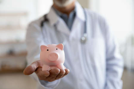 Close up mature doctor therapist gp wearing white coat uniform with stethoscope holding pink piggy bank, healthcare money savings, medical insurance concept, hospital budget and accountingの写真素材
