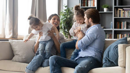 Joyful carefree parents playing with adorable little kids, resting on comfortable couch together at home. Positive happy different generations family having fun with preschool baby daughters indoors.の写真素材