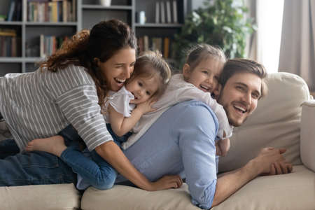 Smiling bonding full family lying on comfortable couch, enjoying playtime together at home. Happy affectionate couple parents spending lazy weekend holiday time with little preschool baby daughters.の写真素材