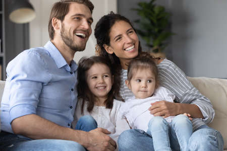 Smiling young married couple relaxing on sofa with adorable little children daughters, enjoying...
