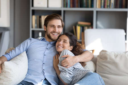 Happy young married couple embracing relaxing on sofa, enjoying sweet tender moment together at home. Smiling affectionate handsome man cuddling attractive woman, showing love feeling excited indoors.の写真素材