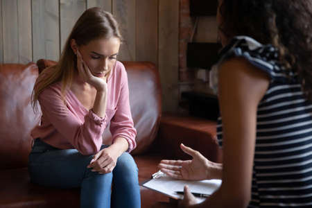 Unhappy depressed young woman listening to psychologist at meeting, African American woman therapist counselor consulting stressed frustrated girl at counselling therapy session, explaining diagnosisの写真素材