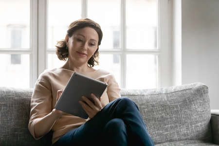 Interested 50s old mature brown-haired woman sitting on cozy sofa, involved in reading online book or media news on computer tablet. Happy senior elderly female shopper watching good in online store.の写真素材
