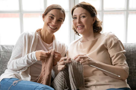 Portrait of smiling young woman resting on sofa with affectionate mature older mommy, holding knitting needles with threads in hands, involved in domestic hobby activity at peaceful weekend time.の写真素材