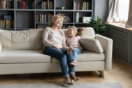 Overjoyed little granddaughter and mature grandmother wearing glasses laughing, sitting on cozy...