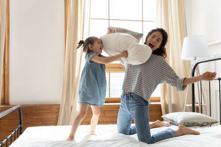 Cheery lively mother and adorable strong daughter holds pillows fighting spend funny time awakened in morning in bedroom. Cozy room interior, crazy kid and mum enjoy playtime activity at home conceptの写真素材