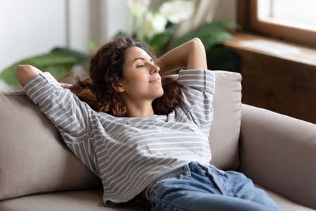 Peaceful young woman relaxing alone in cozy living room leaned on sofa cushion breath fresh air...