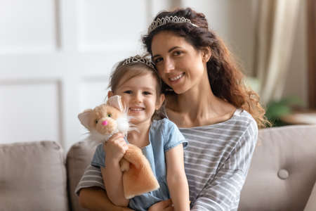 Little daughter hold hand puppet cat fluffy toy sit on mother laps, girls wear crowns smile look at camera. Happy family single mom her kid girl portrait, developing games, having fun playtime conceptの写真素材
