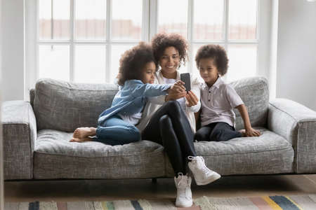 Full length addicted to technology smiling beautiful african american woman showing funny mobile application to happy small daughter ands preschool son, relaxing together on comfortable couch at home.の写真素材