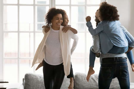 Energetic happy african american couple parents carrying on back joyful small biracial kids, playing catch up at home. Laughing mixed race family of four enjoying domestic weekend activity indoors.の写真素材
