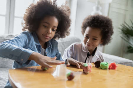 Happy african american older sister and younger brother involved in creating figures with plasticine at home, interested little children siblings developing creativity, improving fine motor skills.の写真素材