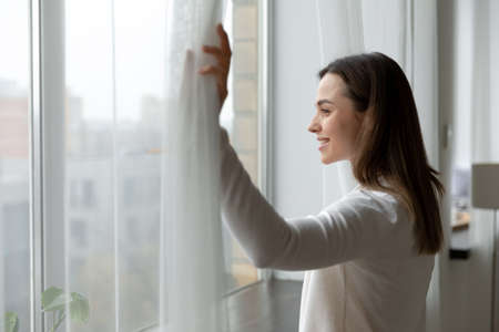 Side view happy attractive woman opening curtains, admiring cityscape at home or hotel room. Smiling...