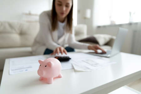 Focus on piggybank with blurred young woman calculating domestic incomes and outcomes on background. Monthly budget management, investment planning, bank loan mortgage credit payments concept.の写真素材