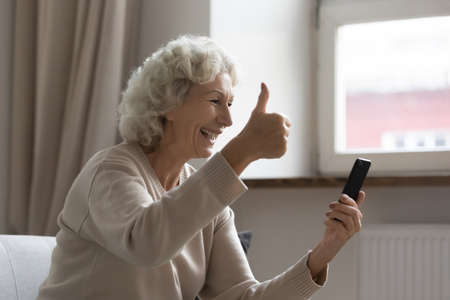 Overjoyed mature woman making video call, showing thumb up, looking at smartphone screen, smiling...