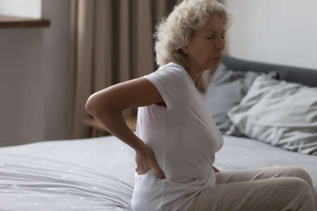Unhappy frustrated middle aged woman touching back rubbing stiff muscles, feeling pain and discomfort after awakening, sitting on bed in bedroom, upset mature female suffering from backacheの写真素材
