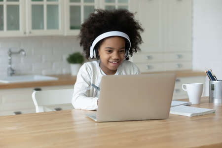Adorable african ethnicity small girl wearing headphones, looking at computer screen. Happy cute...