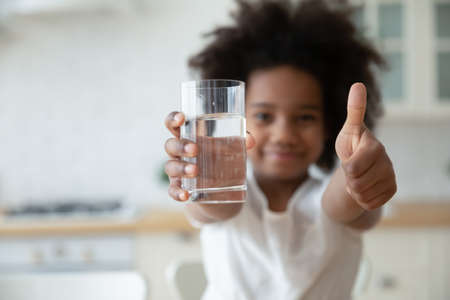Focus on happy small african american girls hands holding glass with fresh pure water and showing thumbs up gesture. Smiling little biracial kid recommending healthcare habit, morning refreshment.の写真素材