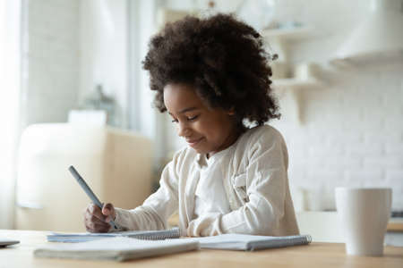 Smiling adorable beautiful african ethnicity little girl sitting at table, writing notes in...