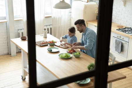 Top view happy young man teaching little adorable child daughter chopping fresh vegetables for vegetarian dinner dish, standing together at wooden countertop, loving family cooking in kitchen.の写真素材