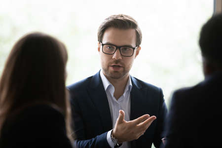 Serious Caucasian young businessman in glasses discuss business ideas with diverse colleagues at team meeting in office. Millennial male boss CEO talk with partners cooperate brainstorm at briefing.の写真素材