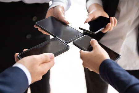 Crop close up of diverse businesspeople hold smartphones communicate via application in office. Colleague coworkers use modern cellphone gadgets at meeting at workplace. Technology, mobile concept.の写真素材