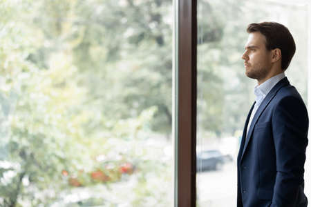 Serious young Caucasian businessman in suit near window in office look in distance pondering or...