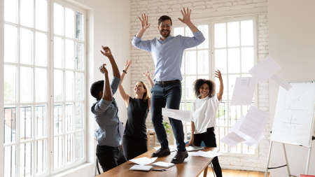 Overjoyed diverse multiracial colleagues have fun at meeting throw papers laugh entertain together, excited multiethnic coworkers dance in office on table celebrate financial company business successの写真素材