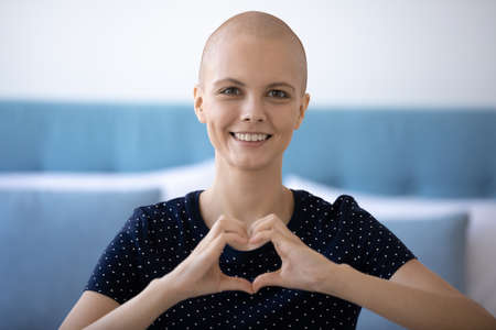 Headshot portrait of happy young hairless woman beat cancer look at camera show heart love gesture...