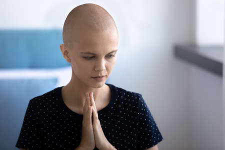 Young hairless sick Caucasian woman suffer from oncology keep hands in prayer pray talk with god at home. Superstitious bald ill female patient struggle with cancer feel religious. Faith concept.の写真素材