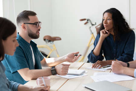 Confident mentor coach young man wearing glasses training diverse employees at corporate meeting,...