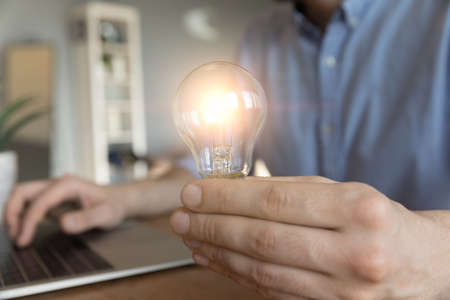 Crop close up of male employee work on computer hold bright light bulb get brilliant business idea. Businessman or worker feel motivated with successful startup project in office. Innovation concept.の写真素材