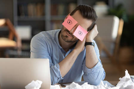 Tired Caucasian male worker fall asleep at workplace in office, have sticker pads on eyes. Exhausted young man take nap doze off sleep at desk, overwhelmed with work. Fatigue, exhaustion concept.の写真素材