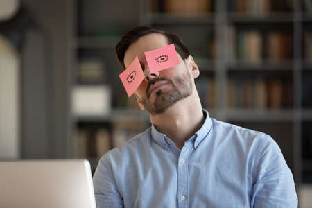 Exhausted young Caucasian man worker have sticker pads on eyes sleeping near computer in office. Tired millennial male employee fall asleep doze off at workplace, feel overwhelmed drained at work.の写真素材