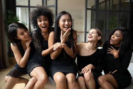 Smiling young chinese 20s pretty girl showing engagement diamond ring to excited diverse female best friends, celebrating hen bachelorette shower party together in black cocktail classy dresses.の写真素材