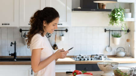 Smiling young Caucasian woman texting messaging on modern cellphone while cooking in kitchen. Happy millennial female use smartphone chatting, preparing tasty healthy breakfast in morning at home.の写真素材