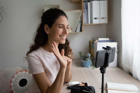 Smiling young Caucasian woman wave greeting with subscribers record video on cellphone camera at home. Happy millennial female vlogger or tutor shoot vlog blog live broadcast on smartphone gadget.の写真素材