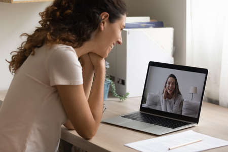 Smiling young woman look at laptop screen talk on video call with girlfriend at home. Happy...
