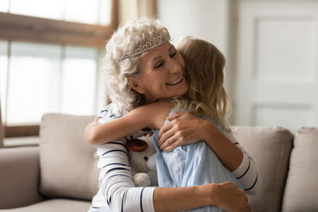 I love you, your majesty granny. Affectionate happy retired grandmother or elderly nanny wearing diadem on silver hair sitting on cozy couch at living room hugging tender little kid girl granddaughterの写真素材