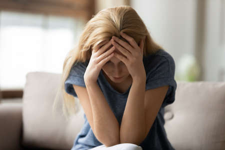 Crazy day. Stressed depressed young adult lady sitting on couch at home with closed eyes hugging...