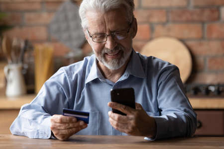 Smiling elderly gray-haired 70s man pay bills online using modern smartphone gadget and credit card....