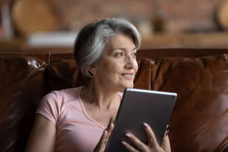 Smiling mature Caucasian grey-haired woman use modern tablet gadget look in distance thinking or dreaming, happy elderly grandmother browse internet read news on pad device relaxing at homeの写真素材