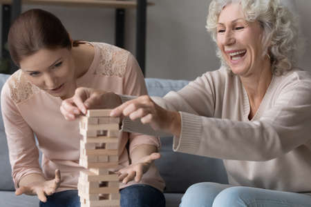 Excited mature woman with adult daughter playing funny stack and crash board game, young female with elderly mother having fun, building tower from wooden blocks, sitting on cozy couch at homeの写真素材