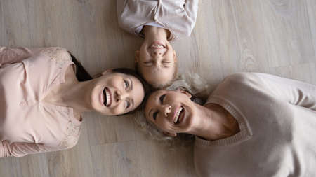 Top view portrait of three generations of overjoyed women lying on warm wooden floor, laughing...