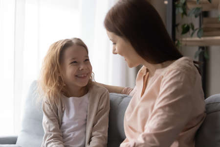 Overjoyed adorable little girl with loving young mother having fun at home, sitting on couch, caring mum hugging excited pretty daughter, chatting, talking, sharing secrets, spending leisure timeの写真素材