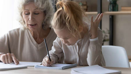 Close up mature grandmother helping unhappy little granddaughter with school homework, difficult tasks, elderly tutor teacher teaching pupil, sitting at desk together, angry girl studying at homeの写真素材