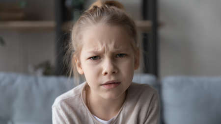 Head shot portrait close up unhappy little girl looking at camera, sitting on couch at home, pretty...