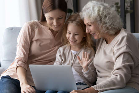Happy little girl with mother and grandmother using laptop together, sitting on couch, looking at...